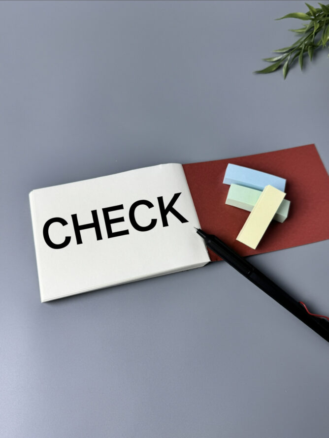 Open notebook page showing the word'CHECK' with pastel sticky notes and a marker on a red folder, on a gray desk; plant in corner above-right.