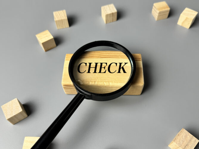 34963877_m | M Virtual(エムバーチャル) - Vライバーマネジメント事務所 Magnifying glass over a wooden block with the word CHECK, surrounded by small wooden cubes on a gray surface.