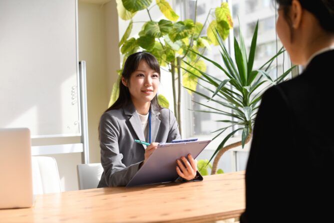Businesswoman in a gray blazer holds a clipboard and talks to a client across a wooden table in a bright office setting with plants in the background.